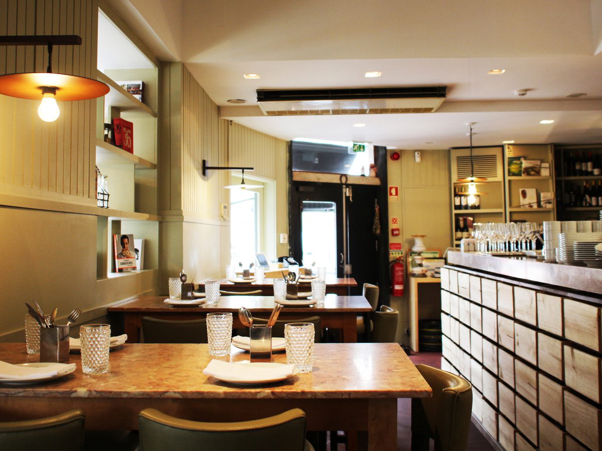 A restaurant interior lit by natural light and industrial pendants on one wall, with tables and a built-in bookshelf to one side and a tiled counter running along the other side, both receding toward the outside door