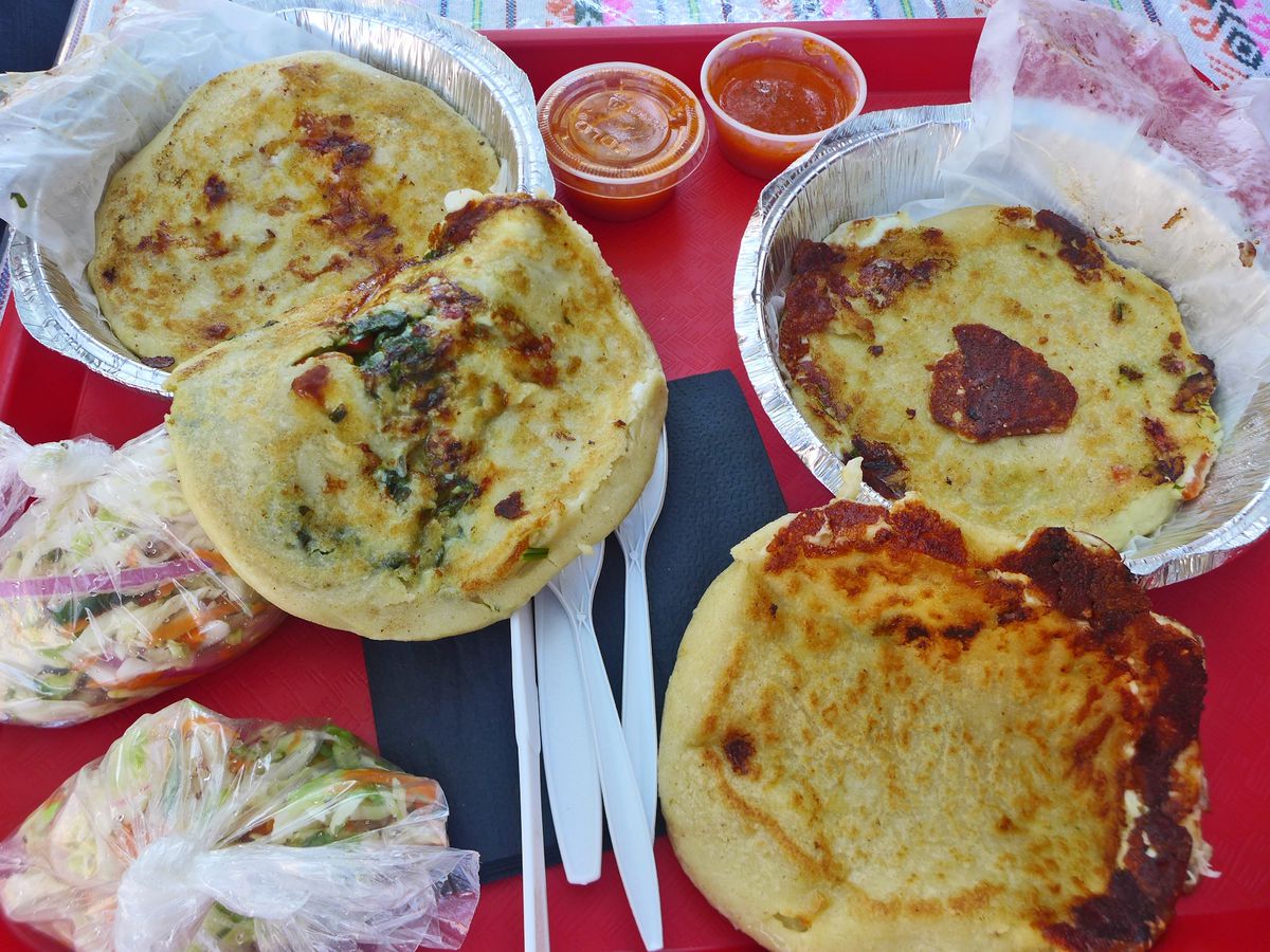 An assortment of outsize papusas, browned stuffed pancakes sometimes broken open to show fillings, on a red plastic tray.