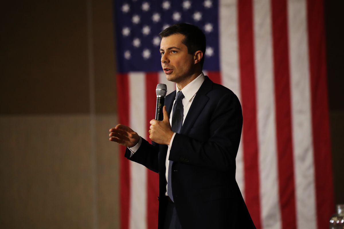 Pete Buttigieg speaks into a microphone in front of an American flag.