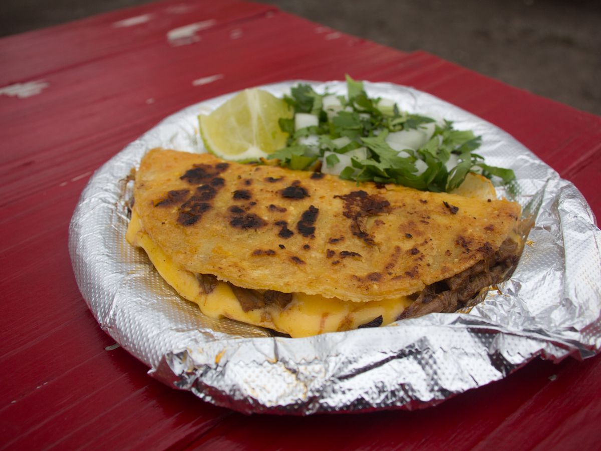 A taco on its side filled with yellow cheese and meat in the foreground, and a pile of cilantro and diced onions and a lime, all on top of an aluminium foil-covered plate on a red table