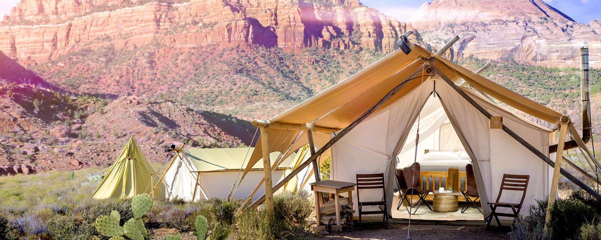 A large tent with its front flaps open is in the foreground. There are chairs and a table in the tent. There are chairs in front of the tent. In the background are mountains in Utah’s Zion National Park.