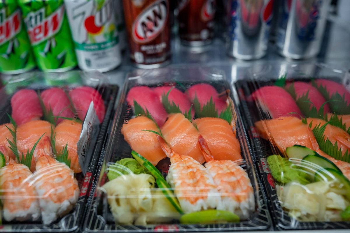 Three grocery-store-style takeout boxes of sushi in a fridge, with shrimp, tuna, and salmon nigiri with wasabi and ginger in each box, with soda containers in the background.
