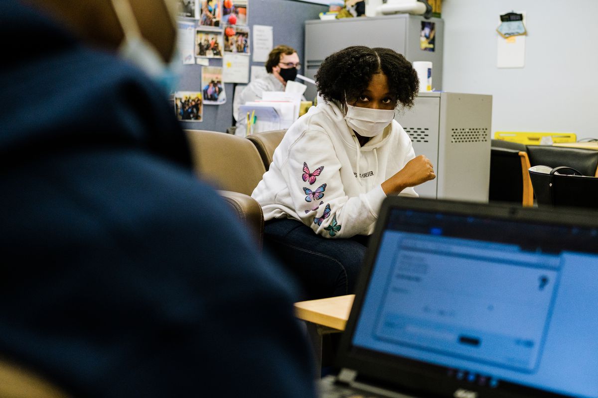 A young woman wearing a protective mask, white hoodie with blue and pink butterflies sits across from a young man wearing a blue hoodie working on a laptop. There is a male teacher sitting behind a desk in the background.