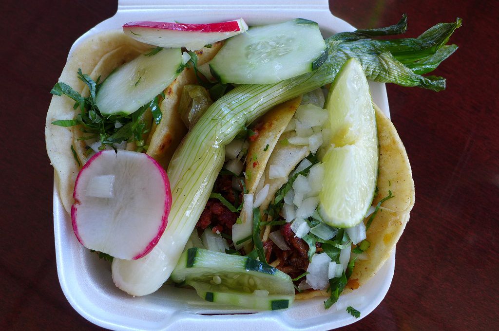 An overhead photograph of a trio of tripe tacos tucked into a takeout container with green onion.
