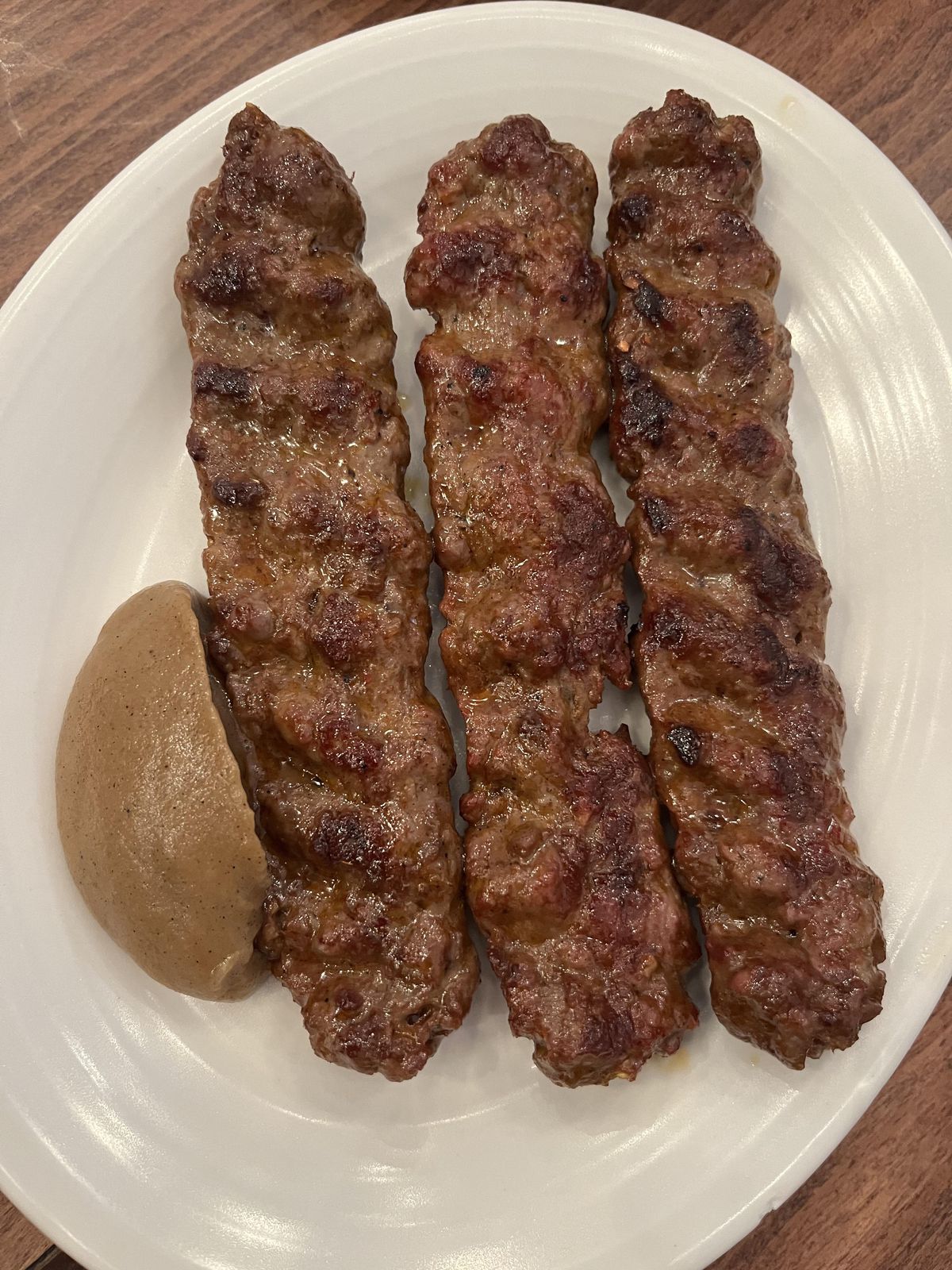 A dollop of grilled apple sauce next to three brown grilled kofte on a white plate, on top of a wooden table.