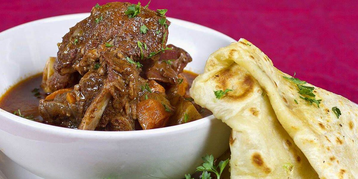 Hunks of goat meat stick out from a bowl of stew along with chunks of potato and carrot, with folded roti leaning against the side of the bowl on a neutral background.