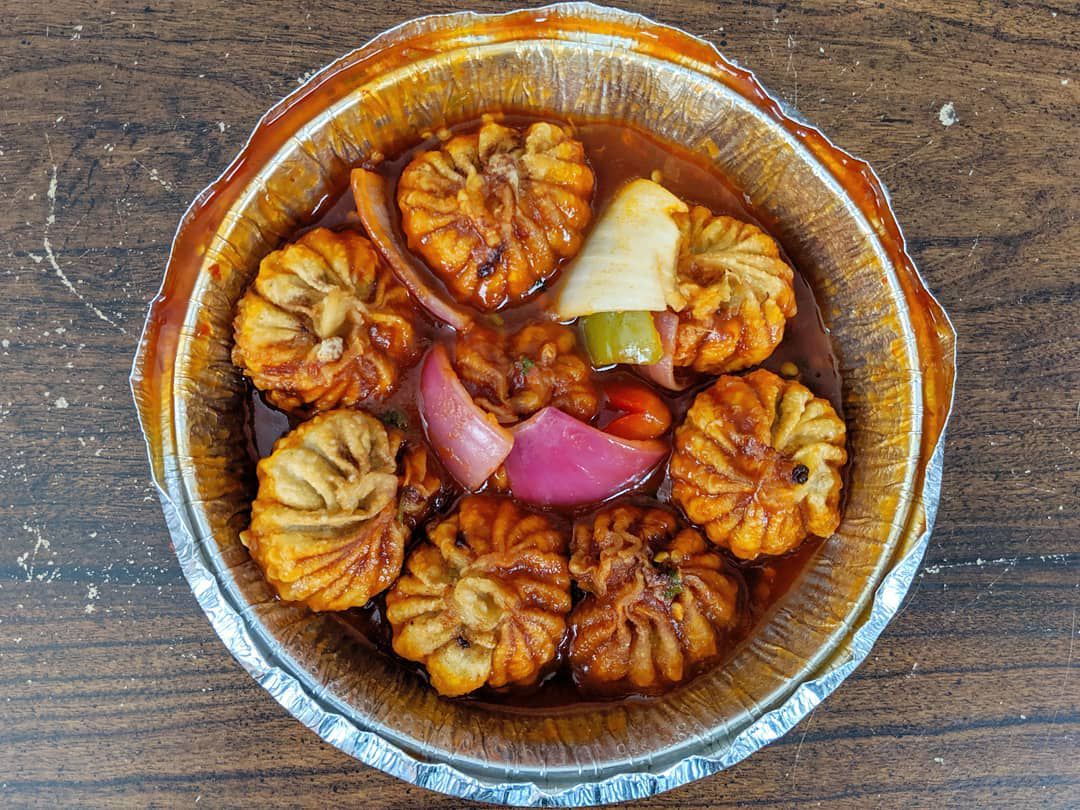 In an overhead view, a round foil takeout container sits on a wooden table, filled with eight fried momo in a red sauce with slices of pepper and onions.