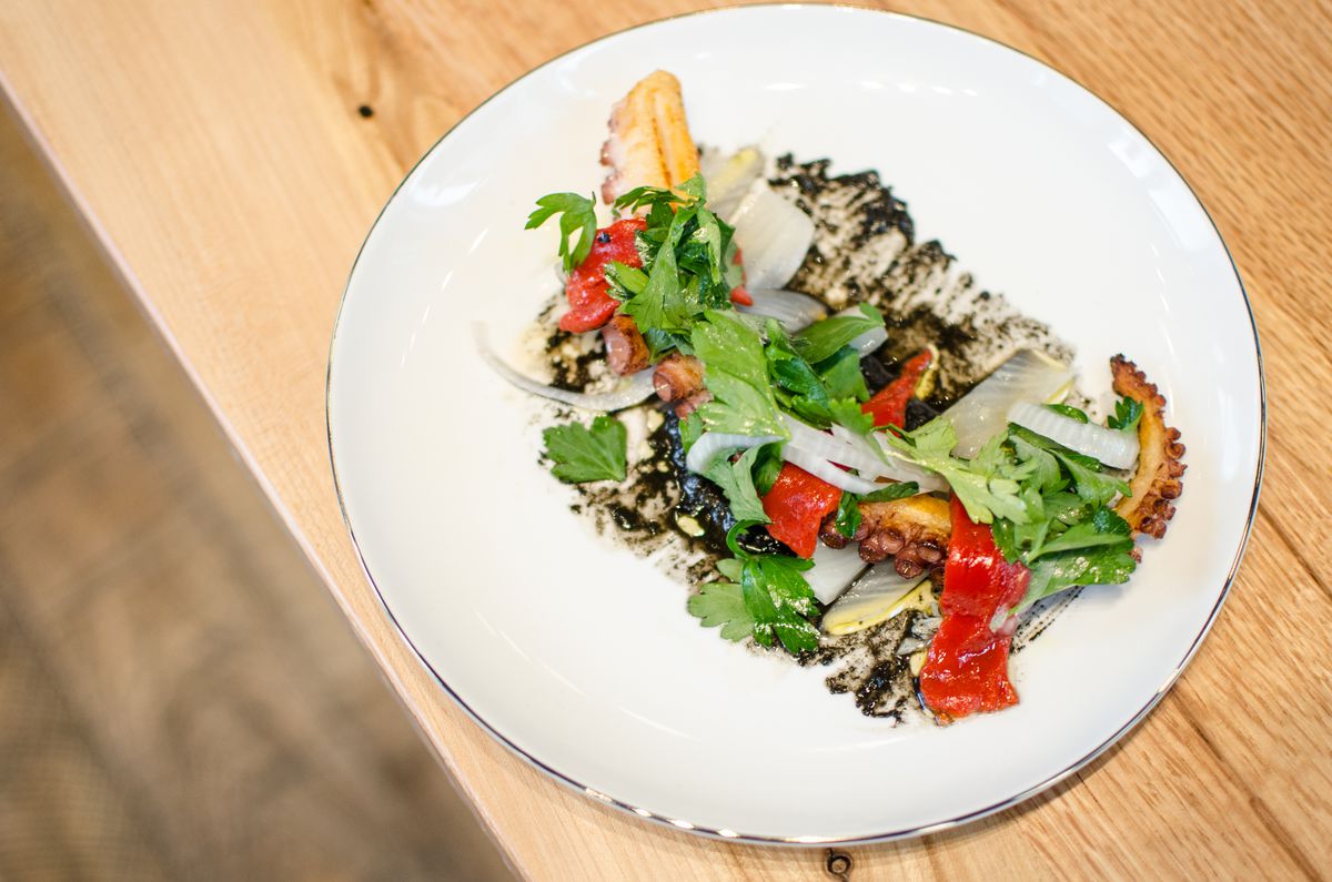 Overhead shot of a charred octopus tentacle on a white plate, garnished with a black sauce, pickled fennel, and bright red peppers. It’s on a light wooden table; a wooden floor is visible underneath.