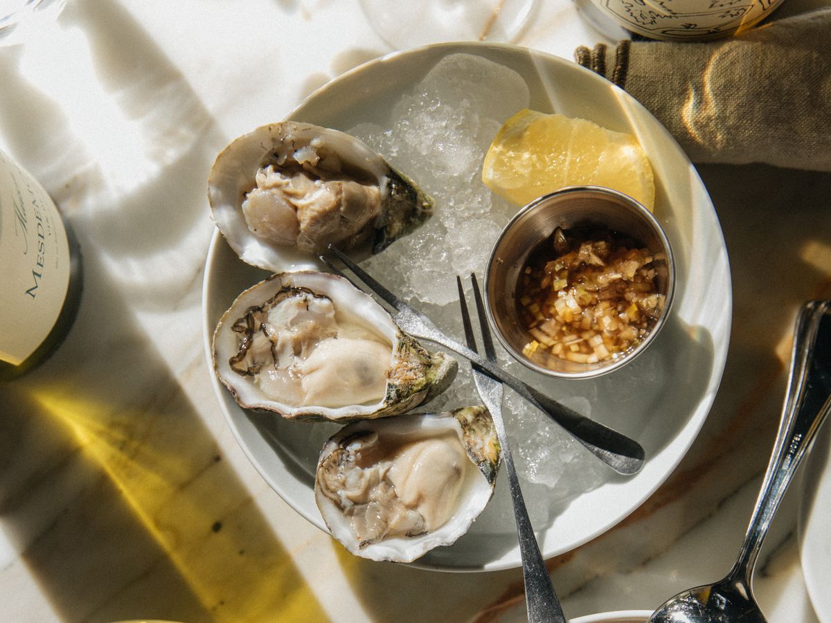 Oysters on ice with horseradish and lemon, beside other dishes and a plate of toast, on a sun-lit table. 