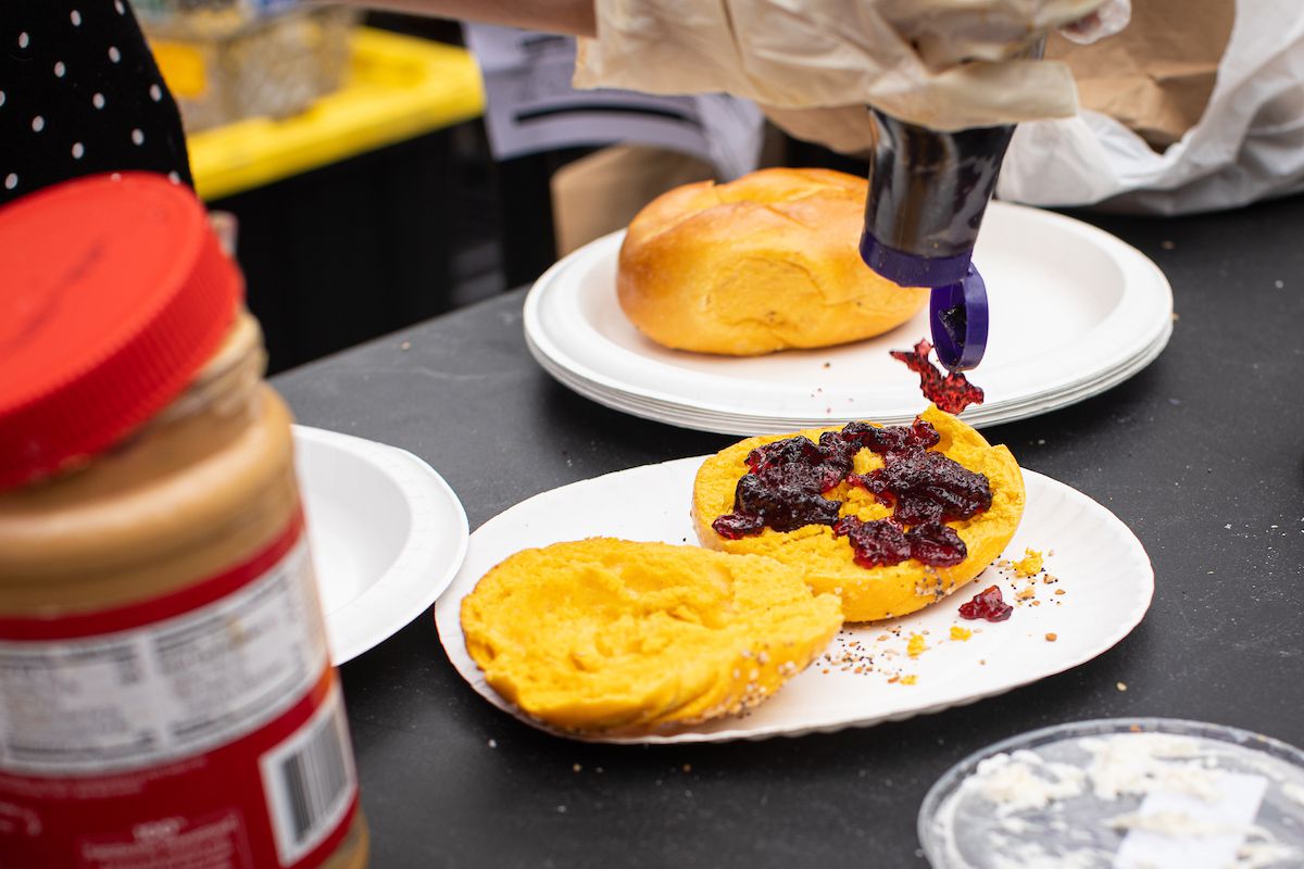 A pair of hands squeeze a bottle of purple jam onto an open-faced egg bagel