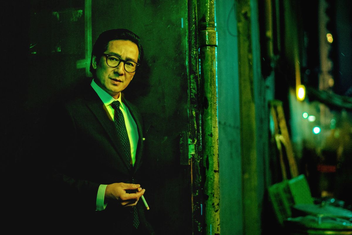 In an image that looks like it’s from a Wong Kar Wai movie, a man in a dashing suit stands and smokes.