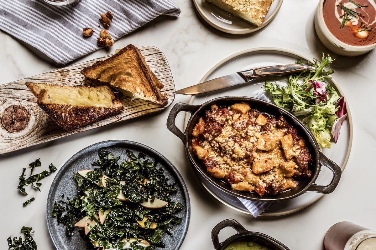 A table is covered in various dishes, including a kale salad, a croissant bread grilled cheese, and gnocchi in a Sunday sugo