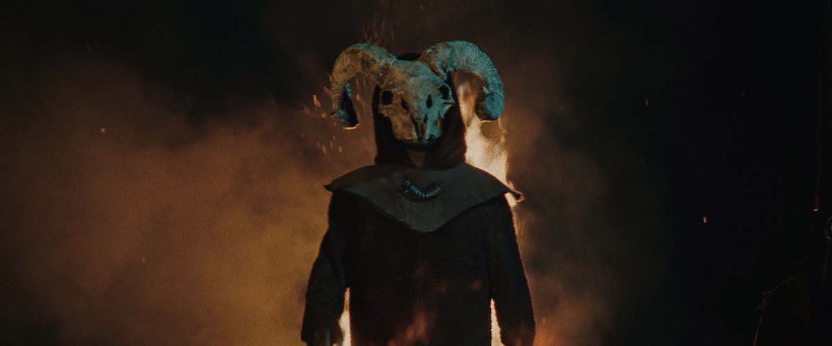 A cloaked figure, wearing the skull of a ram as a mask, stands in front of a flaming backdrop.