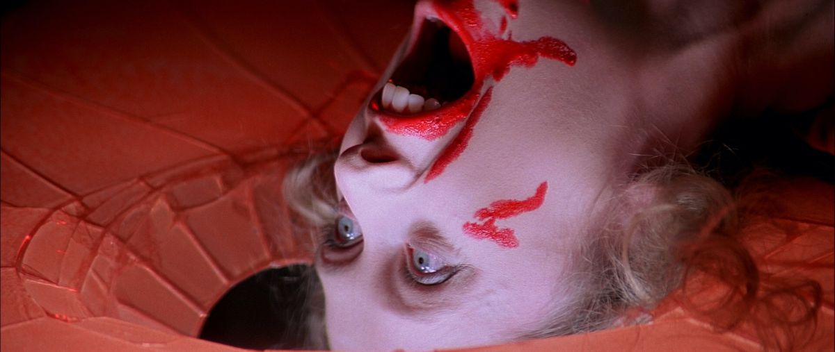 A dead woman lying with her head on a shattered floor, neon-red blood around her mouth, her head thrown back in a final scream, in 1977’s Suspiria