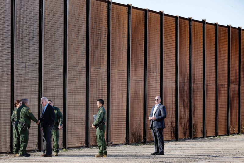 Biden speaks with people in uniform in front of a tall, grim-looking wall.