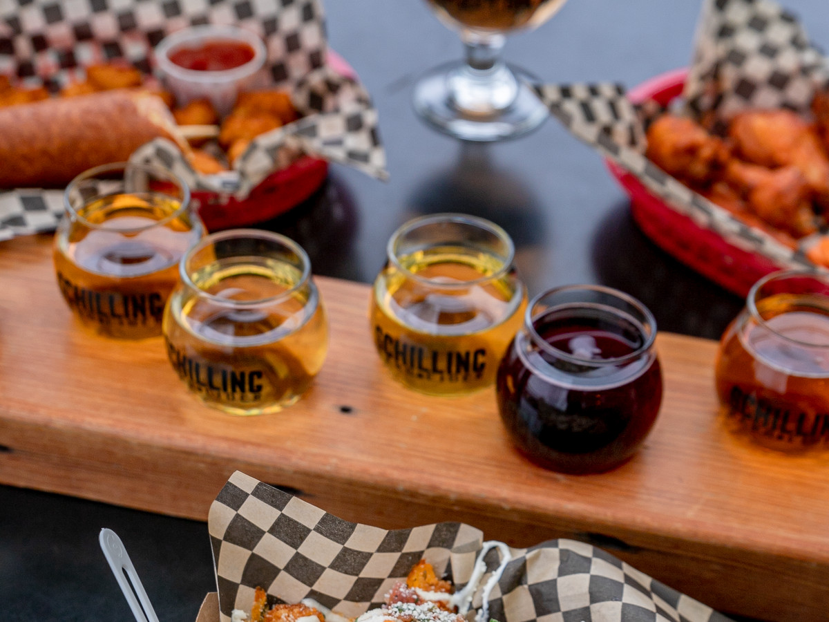 A flight of ciders sits on a wooden tray between baskets of food.