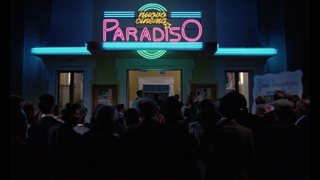 An exterior shot of a crowd huddled in front of the unveiling of the neon sign of the Cinema Paradiso theater.