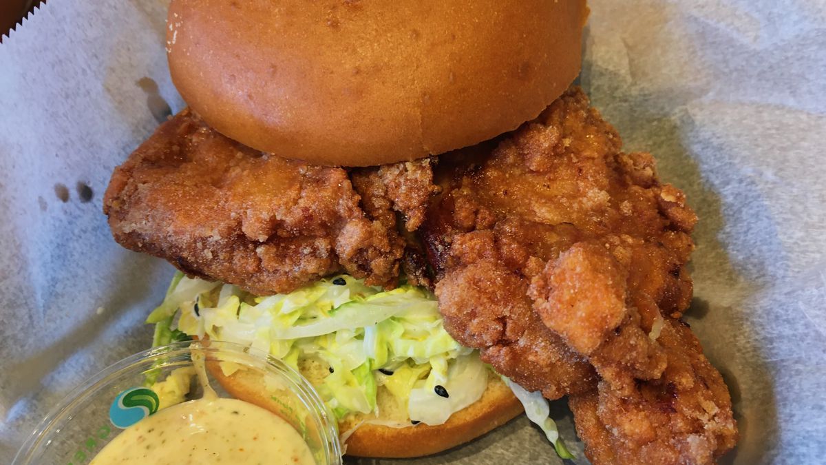 A Japanese karaage fried chicken sandwich on a brioche bun with a citrus slaw and white, creamy sauce in a plastic cup on the side. 