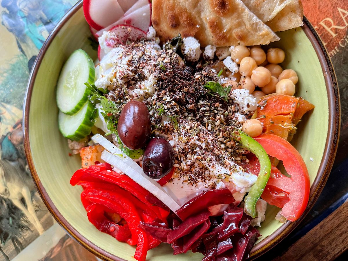 A green bowl filled with colorful veggies, chickpeas, poached eggs, a dusting of za’atar, and pita on the side.