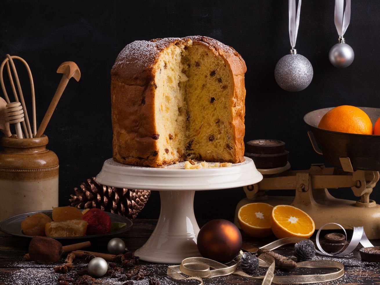 Three quarters of a panettone, with the fluffy yellow and fruit-studded inside of the loaf facing the camera, on a white cake plate, next to a bowl of oranges, Christmas ornaments, and cooking tools.