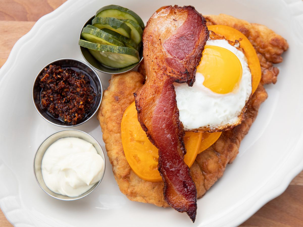 A dish of fried pork with yellow tomatoes, a fried egg, and a slice of bacon layered on top. Three cups of condiments are on the side. The food is set on a white plate.