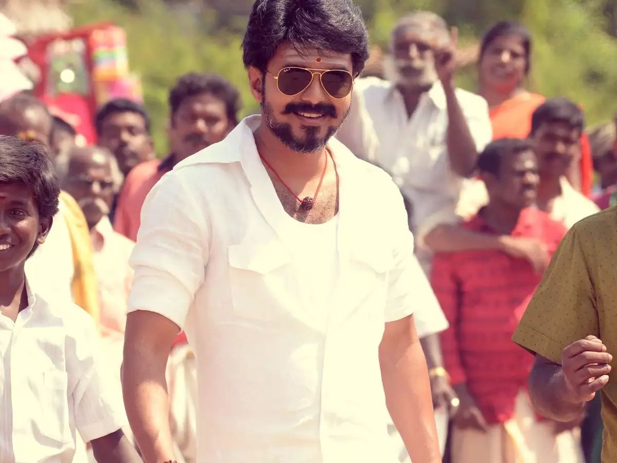 Vijay wears a short-sleeve button-up white shirt, with a white undershirt and a necklace. He also wears sunglasses and has a goatee. His forehead sports a tilak and he is surrounded by a crowd, in Mersal.