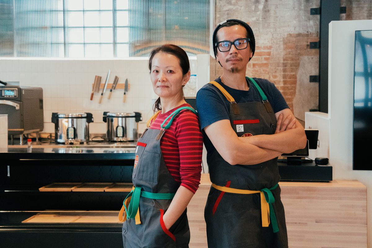 Lina Goh and chef John Ng stand wearing matching gray aprons in a kitchen space with an exposed brick wall. 