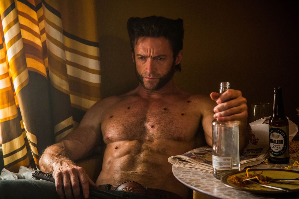 Wolverine, played by Hugh Jackman, sits in a chair. His expression is clearly irritated as he sits shirtless, one hand gripping a bottle of what’s probably alcohol.