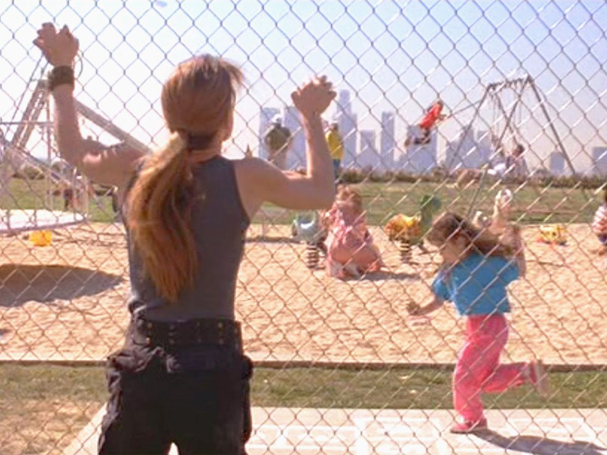 Sarah Connor grips a chain link fence as she stares out at a Los Angeles playground.