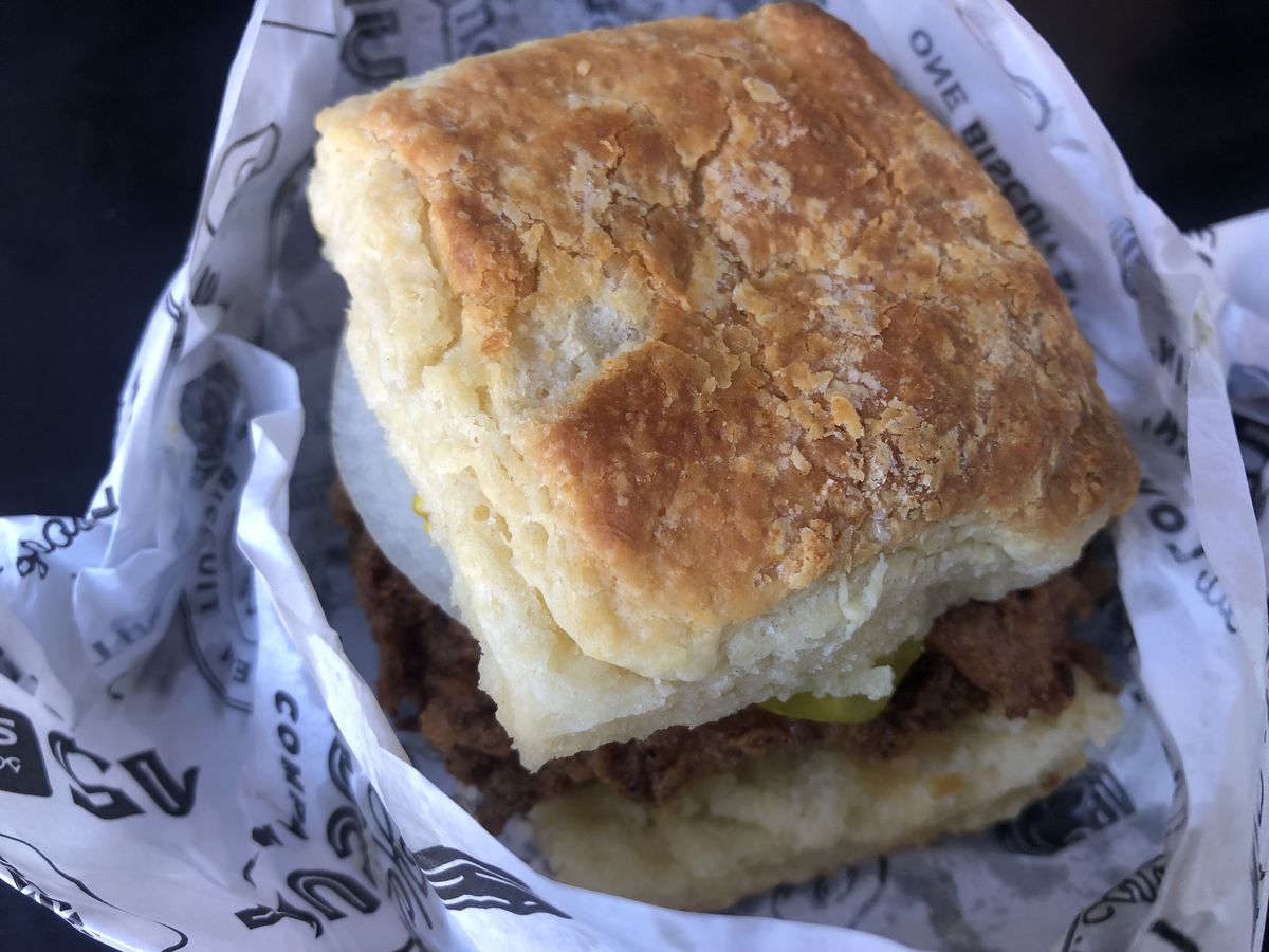 A close-up photo of an unwrapped biscuit sandwich stuffed with fried chicken, pickles, and onion slices.