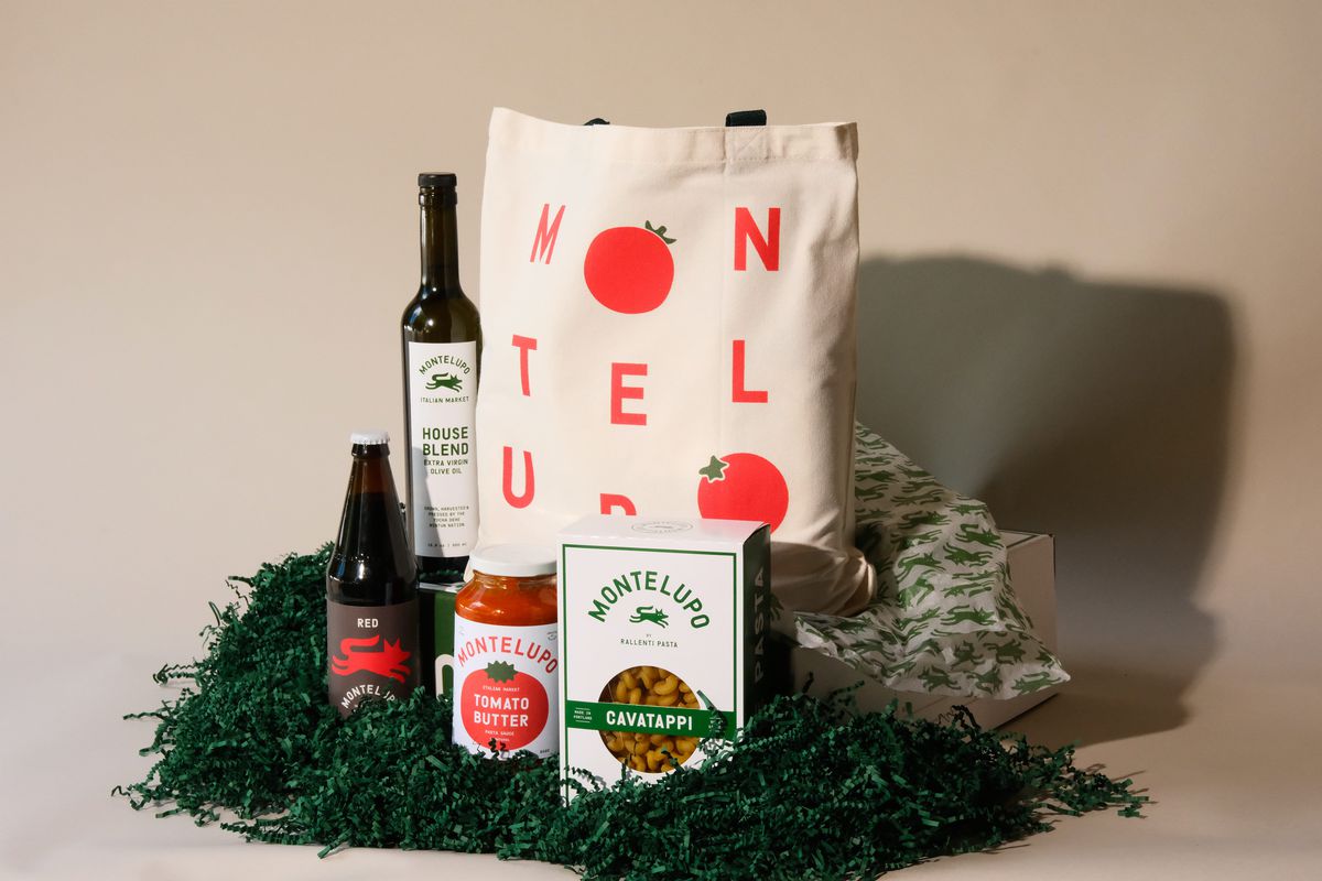 A box of Montelupo cavatappi, a jar of tomato sauce, a bottle of red wine, a bottle of olive oil and a red and white tote bag rest on a bed of green confetti.