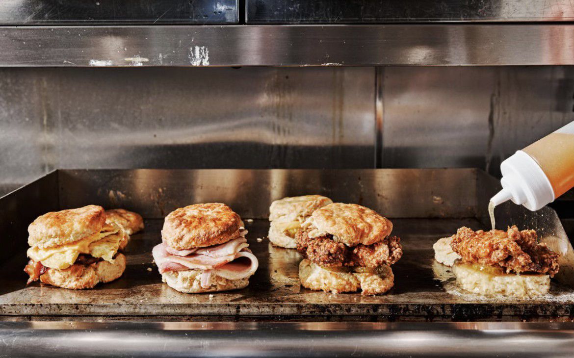 Four biscuits, one with bacon, egg, and cheese, one with ham and cheese, and two with fried chicken, sit on the griddle at Bomb Biscuit in Atlanta.