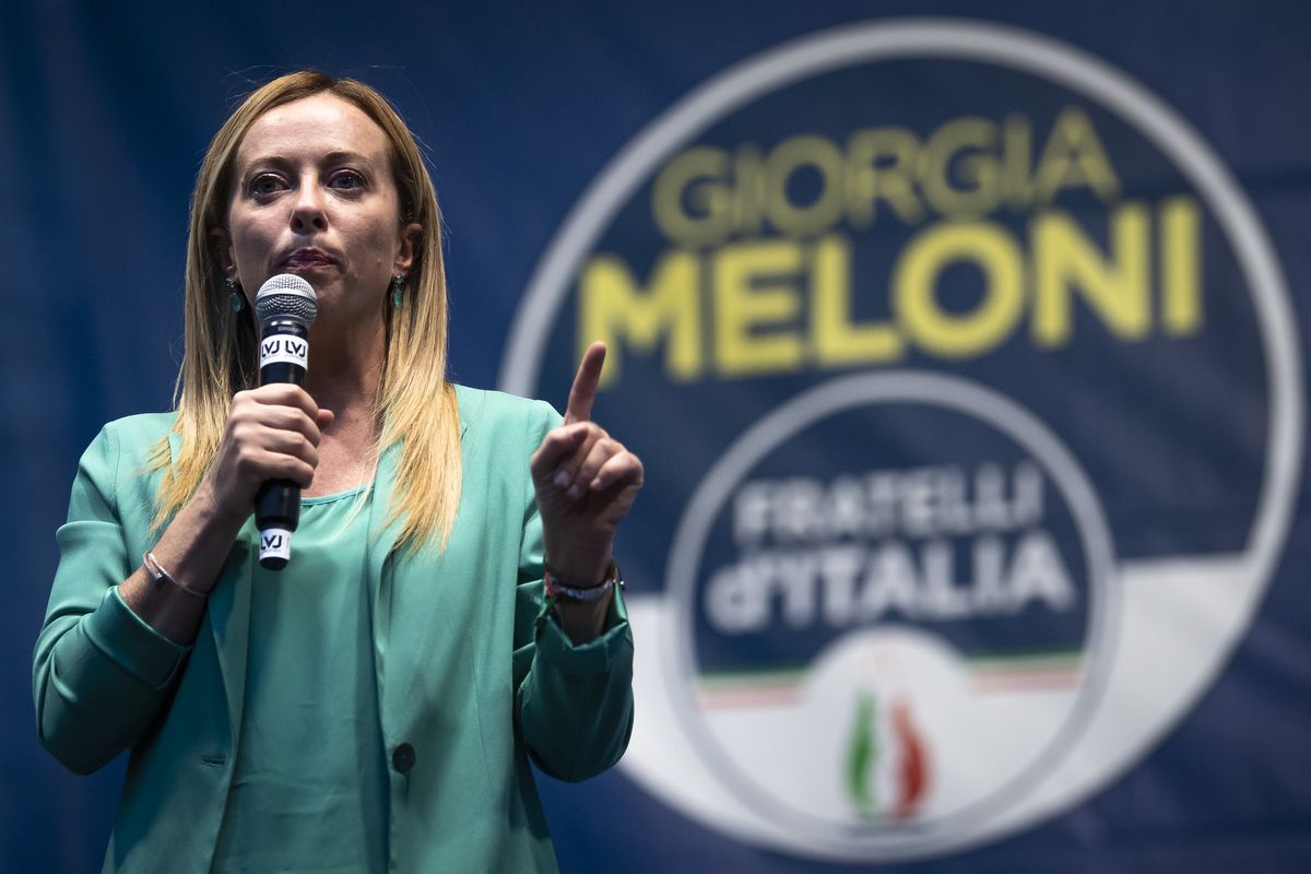 Italian politician Giorgia Meloni gestures while holding a microphone and standing in front of a banner with her name on it.
