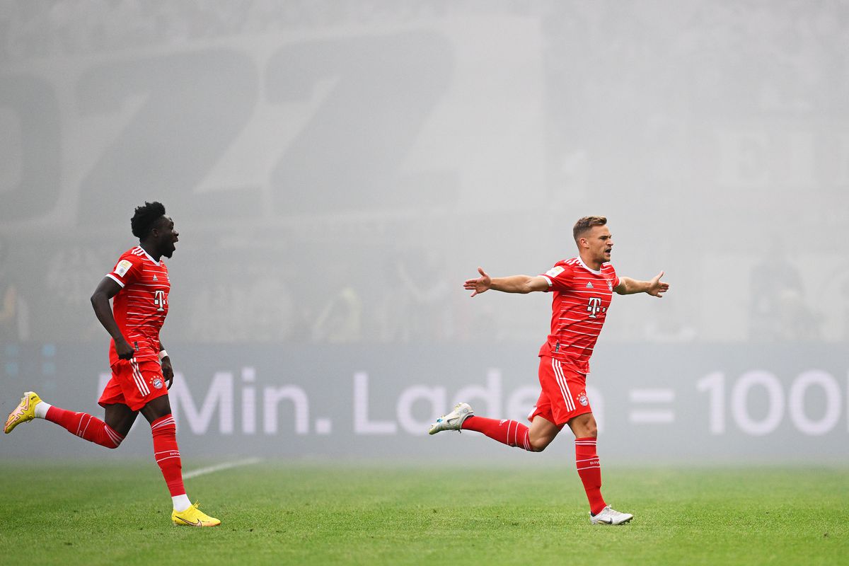 Joshua Kimmich spreads his arms in the air and runs to the sideline to celebrate his goal against Eintracht Frankfurt on opening day, Alphonso Davies trailing behind him, amid a smoke-filled background.