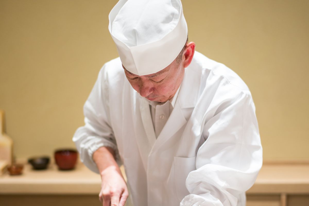 Eiji Ichimura wearing chef’s whites and a white hat behind a sushi counter.