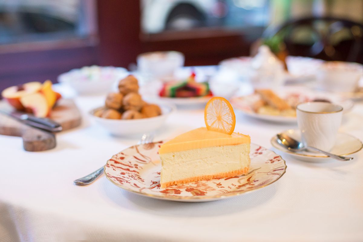 A piece of lemon cheesecake, garnished with a dried lemon, sits on a white china plate with red patterns. Other dishes are on the table in the relief.