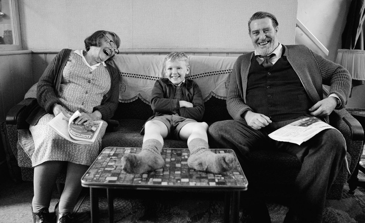 A young boy sits on a sofa between two older people, his grandparents. The image is in black and white.