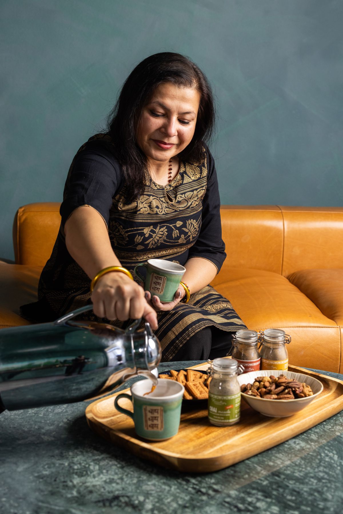 A woman sits on a brown leather couch and pours chai from a sterling silver pot into a green mug that sits on a tray with various snacks.