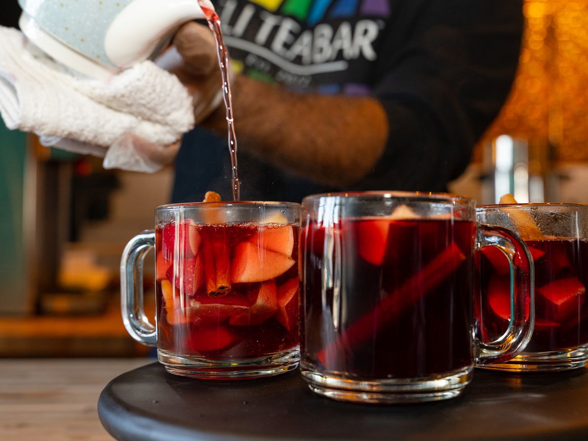 A person pours tea out of a teapot into one of three glass mugs filled with red tea, fruit, and cinnamon sticks.