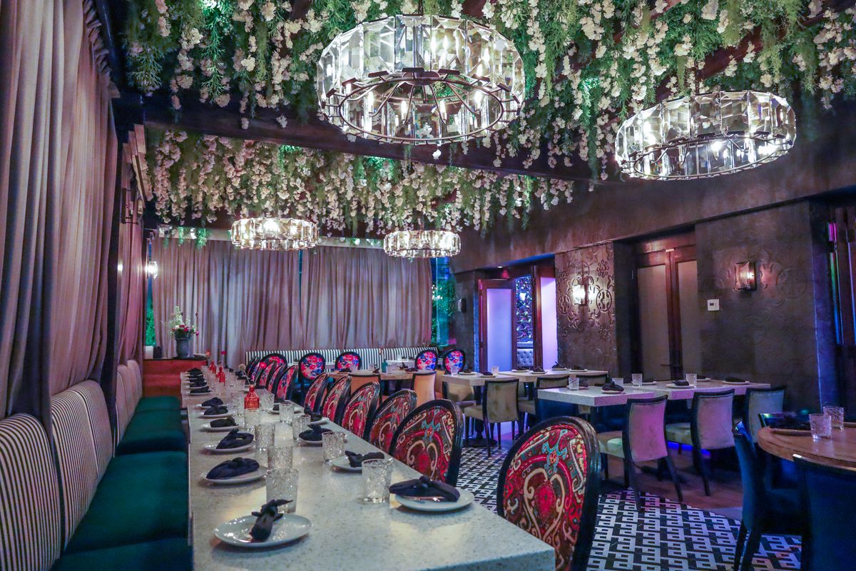 Dining tables surrounded in pink curtains with four chandeliers and strings of fake flowers hanging overhead.