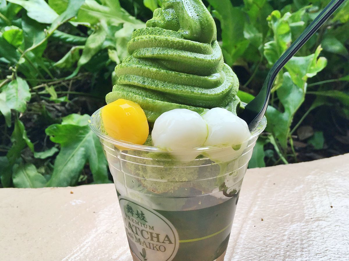 A plastic cup filled with mochi, cake, and other fillings, topped with a large sculpted dollop of matcha soft serve with a plastic spoon sticking out.