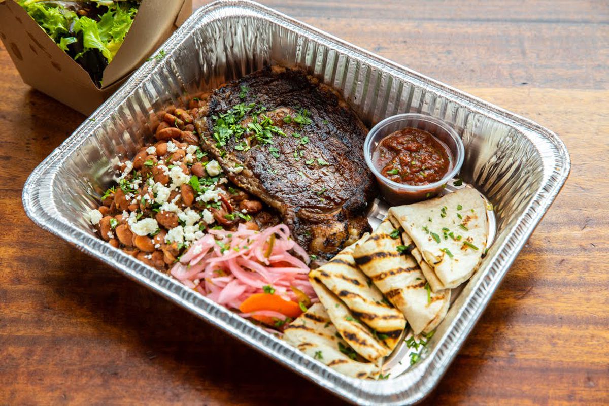A steak, beans, tortillas, and pickled onions sit in a takeout container on a wooden table.