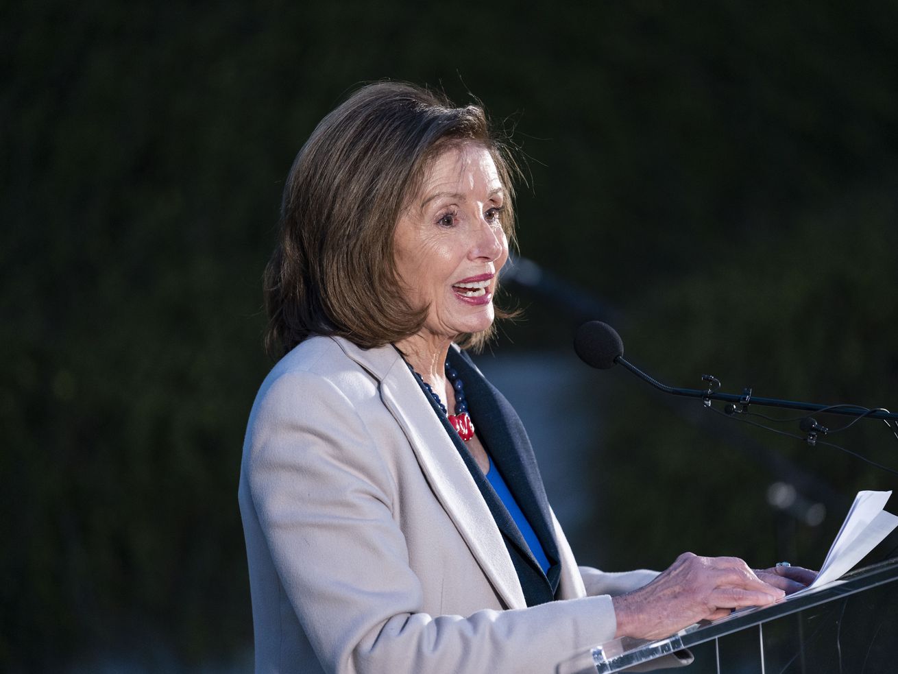 Pelosi, in a pale blue suit, is seen in profile against a dark background, speaking into a microphone.