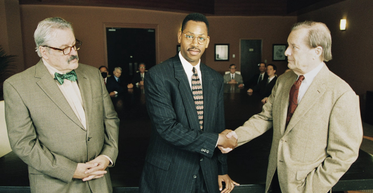 A still of two white men flanking a Black man, who is shaking on of their hands and is facing the camera