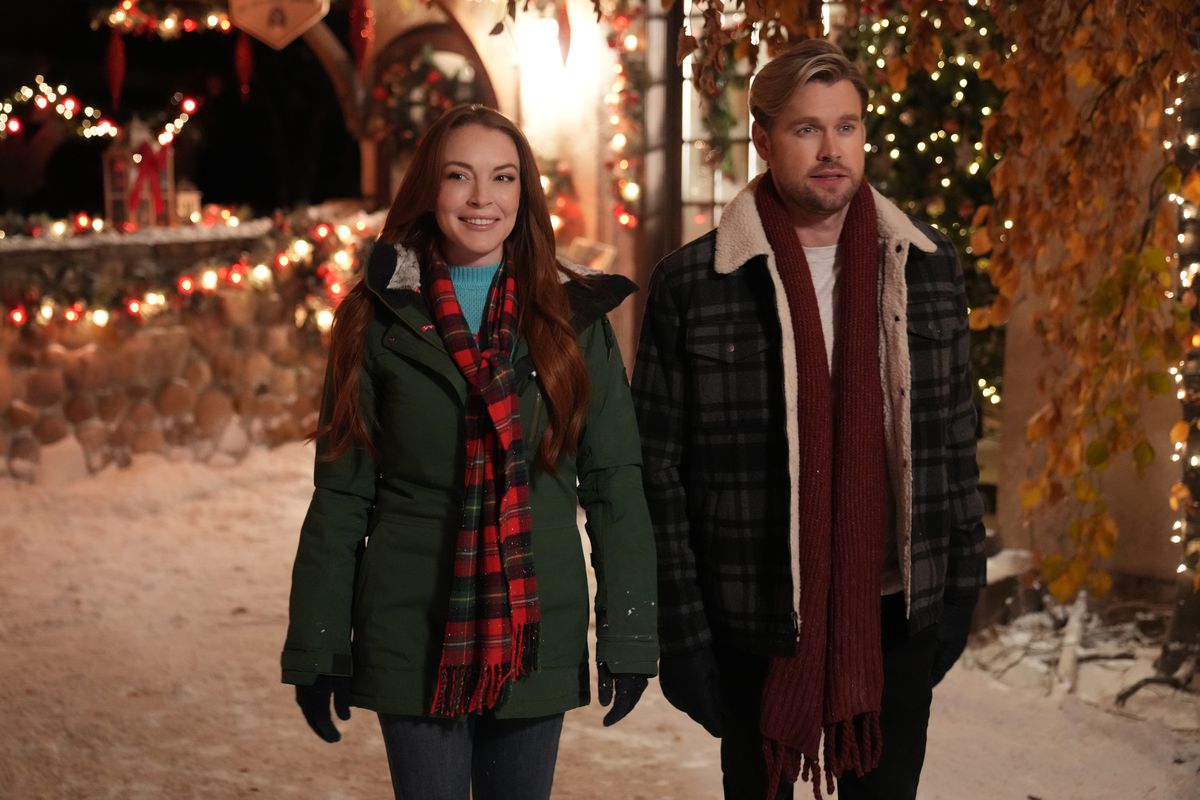 A woman in an olive green winter jacket and red checkered scarf (Lindsay Lohan) walks alongside a man (Chord Overstreet) outside a house decorated in Christmas lights.