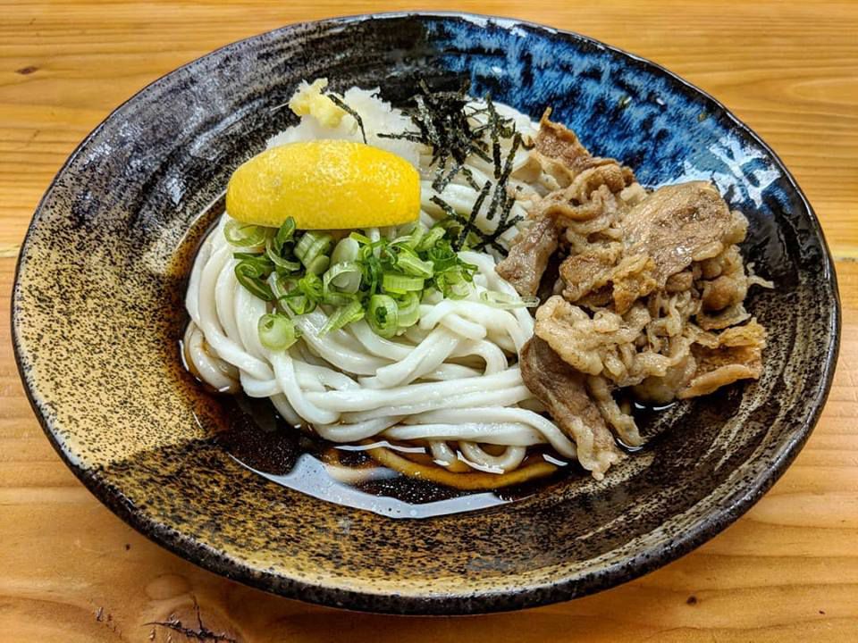 A black bowl that has an artistic, handmade look is full of udon and thinly sliced beef in a thin brown broth. The udon is topped with chopped scallions, slivers of seaweed, and a lemon wedge. The bowl sits on a light wooden table.