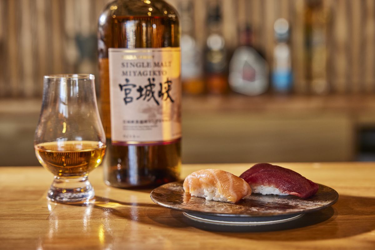 A bottle of Japanese whiskey sits in the middle of a frame, on a table. Next to it is a glass that is a quarter full. To the right is a small plate with two pieces of nigiri. 