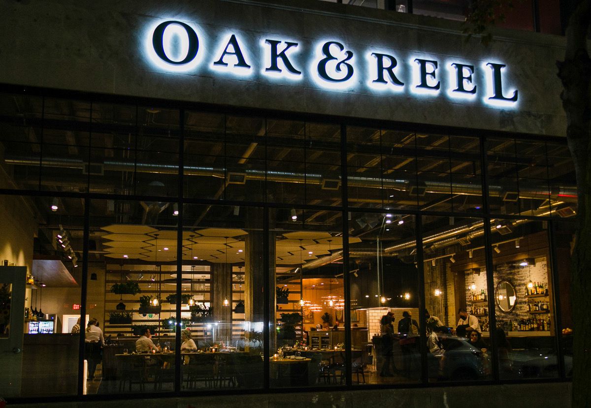 Diners are view through large exterior windows beneath the sign for Oak and Reel at night.