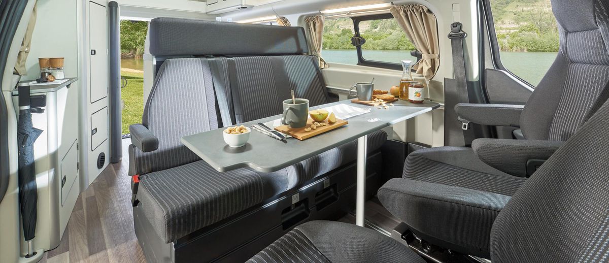 A gray interior of a pop-top camper van comes with seats for five and a table in the middle with food on it. 