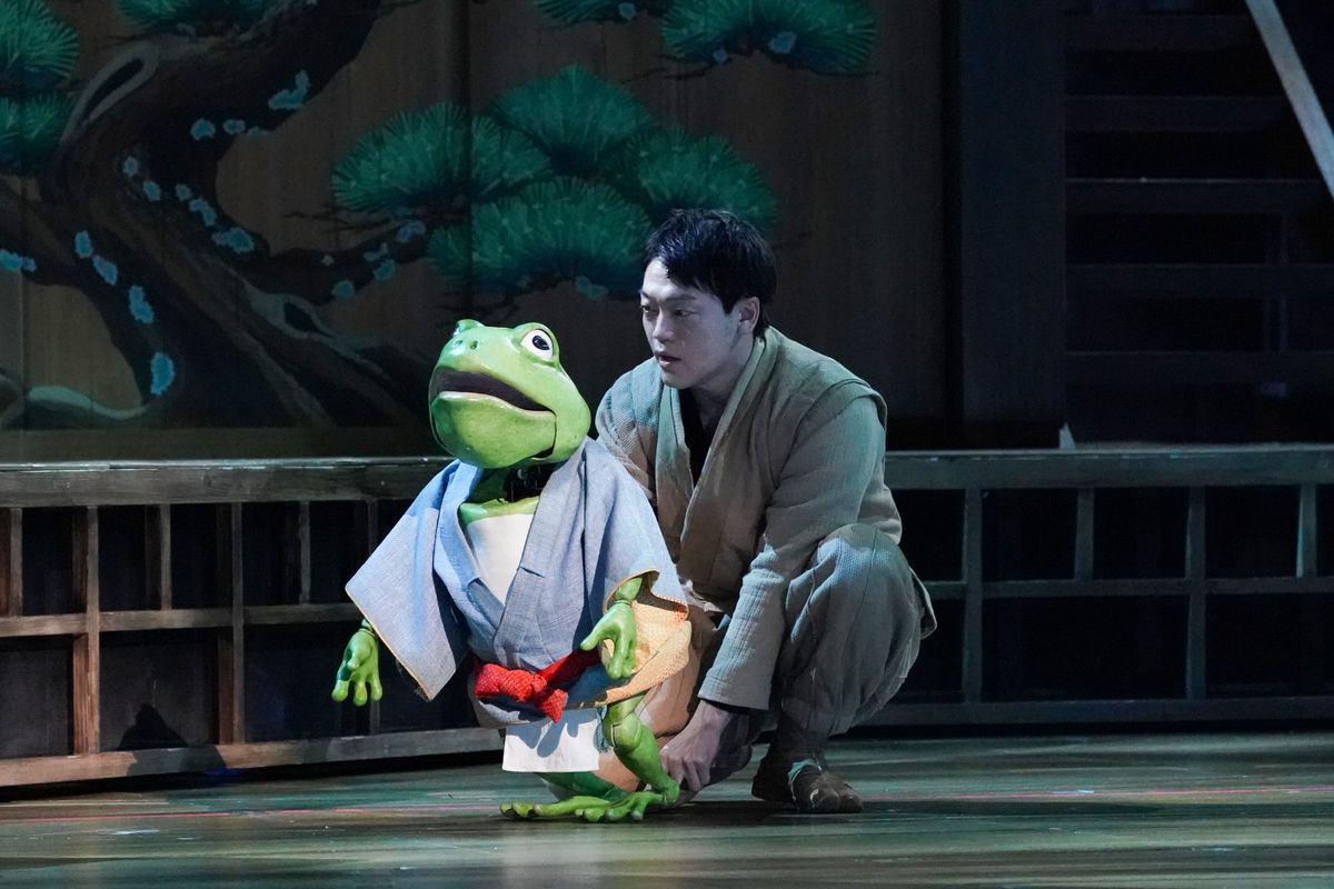 A puppeteer crouches on the stage, operating a frog puppet in Spirited Away: Live on Stage